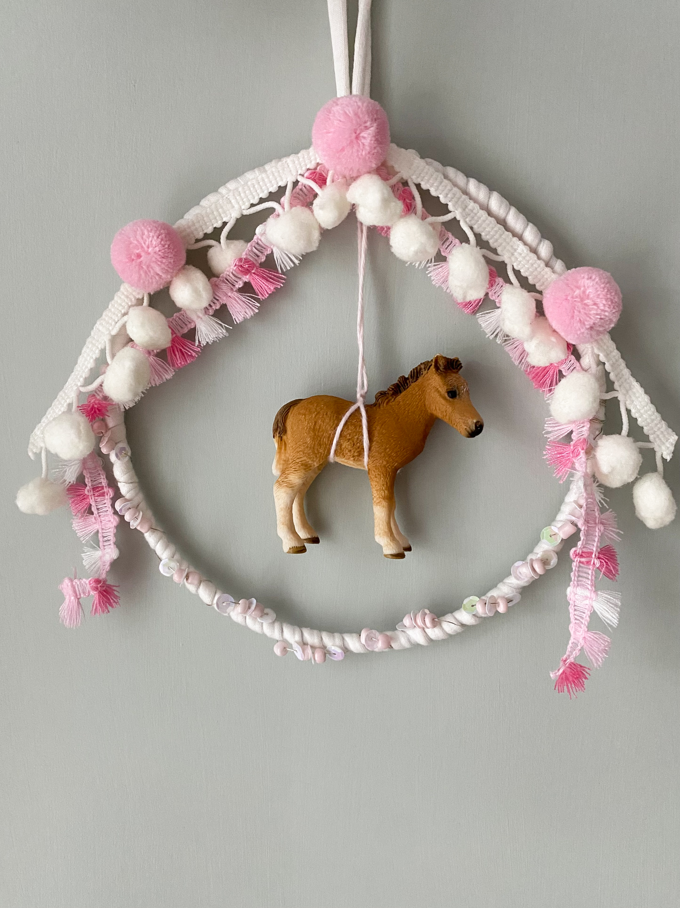 Mobile aus weiß-grauem Makramee, rosa und weiße Pompons, hängendes Spielzeugpferd in Braun, Textilfransen in Rosa und Weiß.