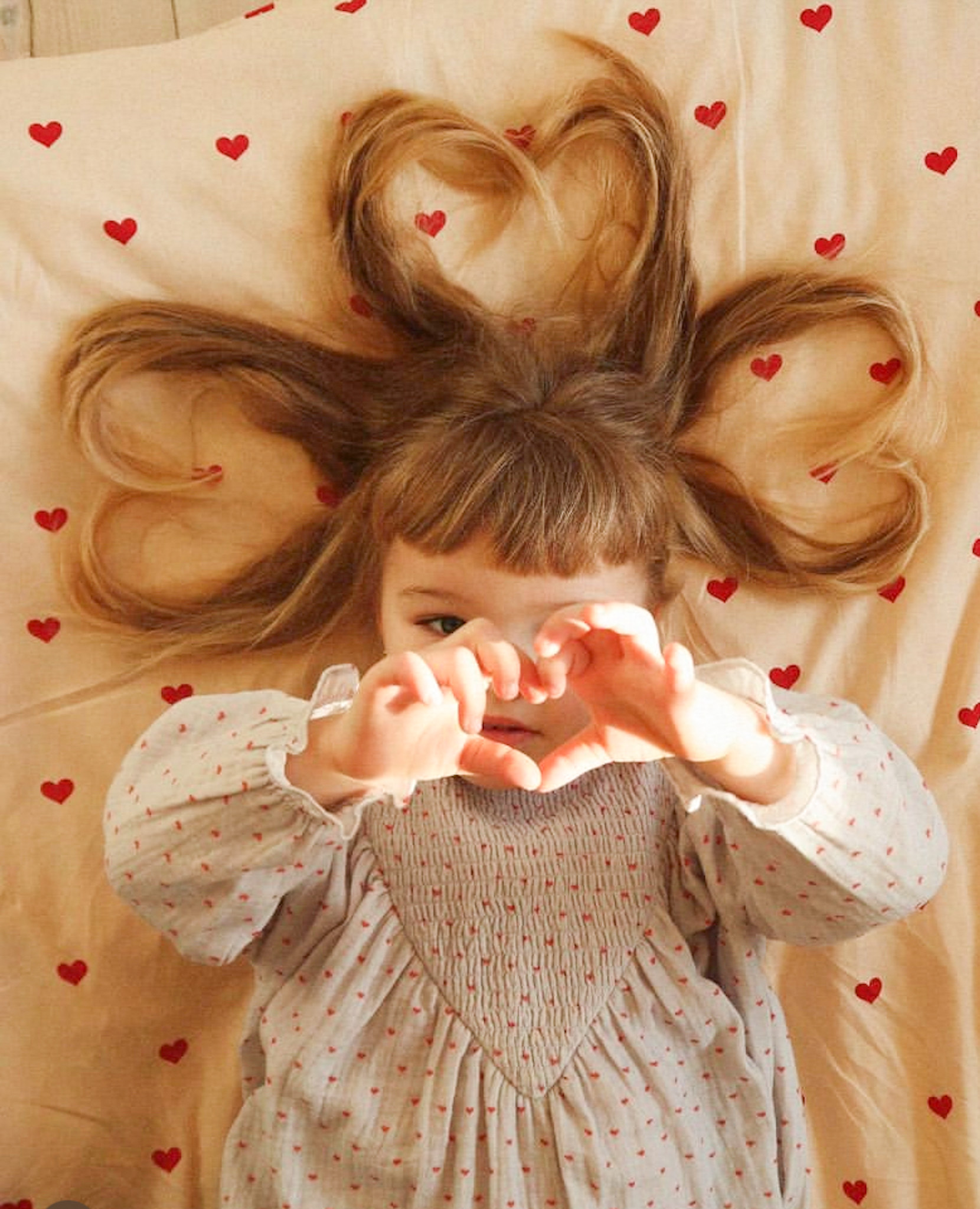 Kleinkind; langes Haar; Herzform-Frisur; beige Bettwäsche; rote Herzmuster; graues Kleid; rote Punkte; Hände formen Herz.