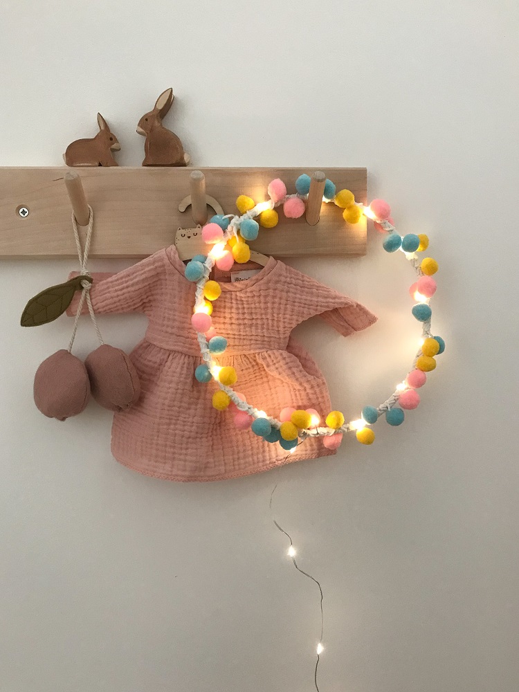 IMG_4947 Holz-Kleiderhaken, zwei Hasenfiguren, rosa Kinderkleid, Pompon-Lichterkette in Blau, Gelb, Rosa.
