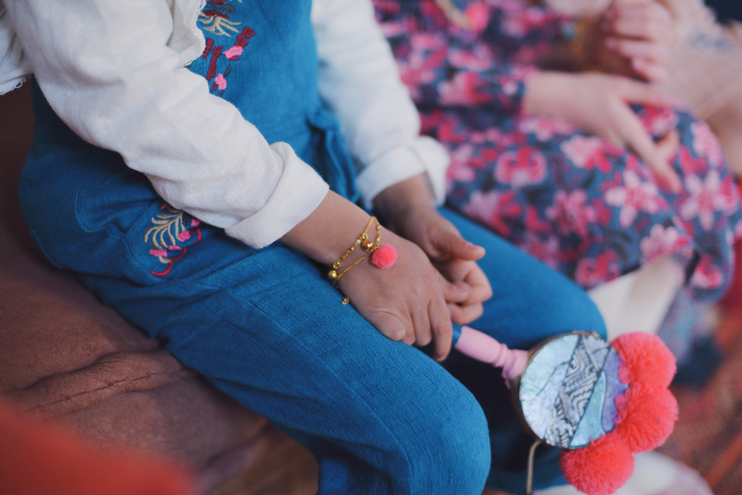 kidsbraceletkamilfluopink2 Kind trägt blaue Hose mit Stickerei, weißes Oberteil; rosa Pompon-Armband.