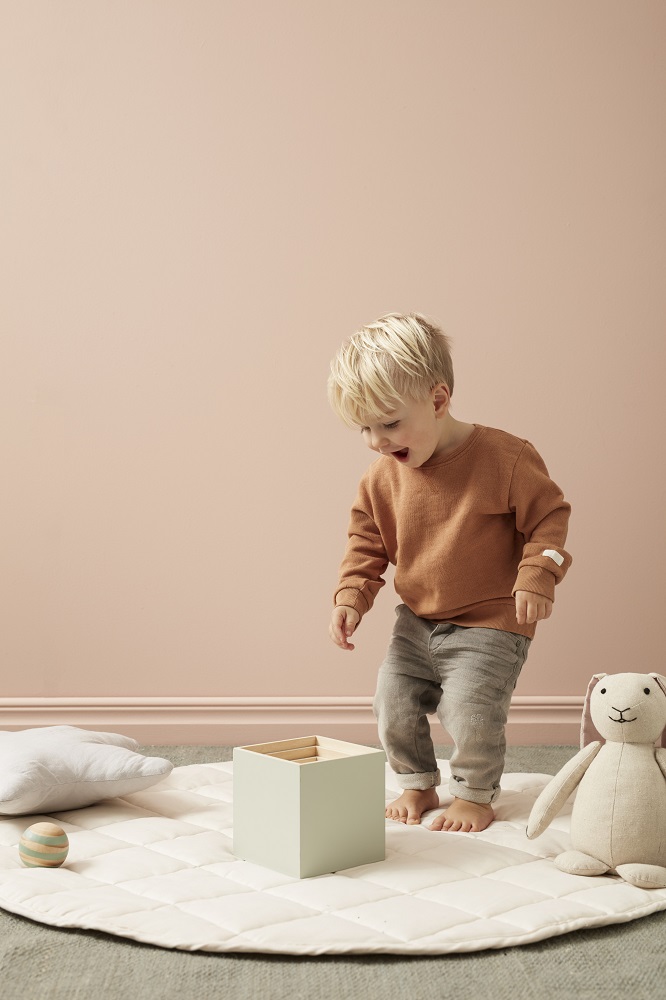 1000483-Play-mat-Beige-1000451-Wooden-Stacking-Cubes-Edvin-EWM_2 Kleinkind, brauner Pullover, graue Hose, hellgrüne Spielbox, weiße gesteppte Spielmatte, Stoffhase, bunte Holzkugel, beiger Hintergrund.