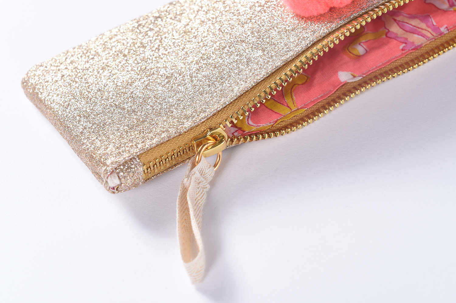 OUSTINA_5 Kosmetiktasche, goldene Glitzeroberfläche, Reißverschluss, pinkes Innenfutter mit Blumenmuster, roter Pompon-Anhänger.