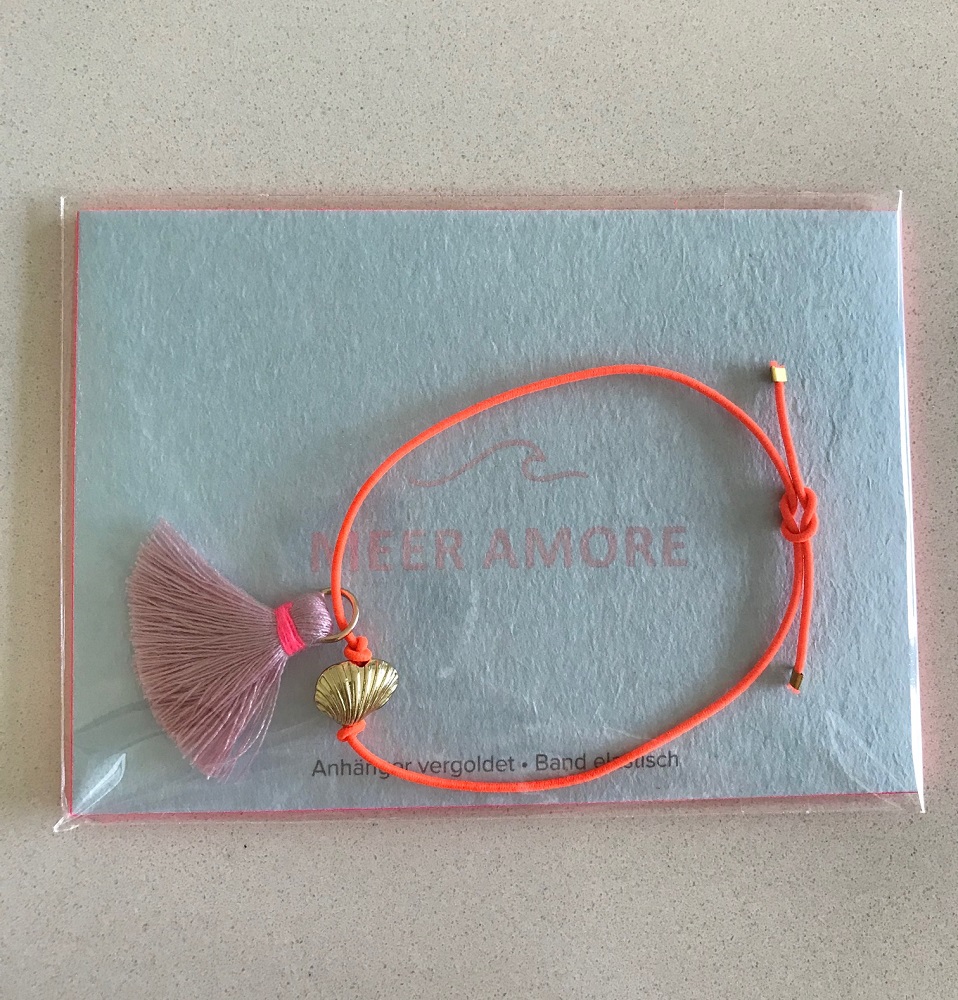 IMG_5906 Armband, orange Band, goldener Muschelanhänger, lila Quaste, transparente Verpackung, Hintergrundkarte mit Aufdruck "Meer Amore".