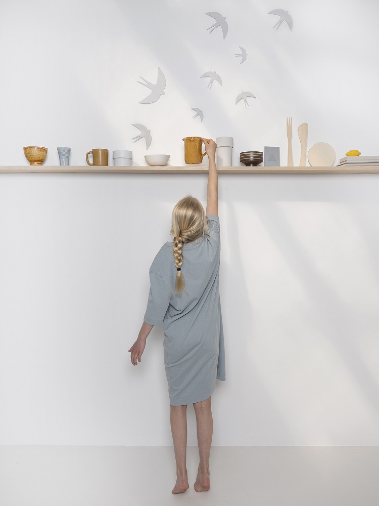 sverm-birds-large-4 Kind in grauem Kleid, Greifen nach gelber Keramikvase auf Holzregal; neutrale Tassen, Schalen, Teller, Holzbesteck; Wanddekoration Schwalben-Silhouetten.