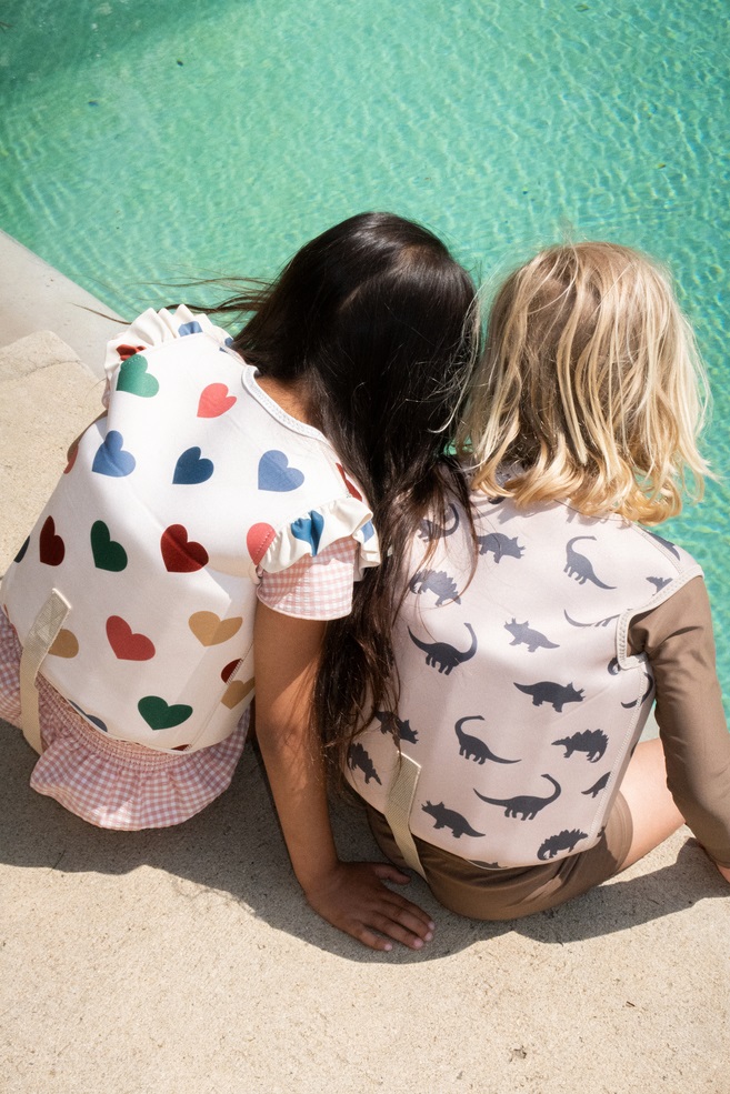 Zwei Kinder-Schwimmwesten; links weiß mit bunten Herzen, rechts beige mit schwarzen Dinosauriern; am Beckenrand sitzend.