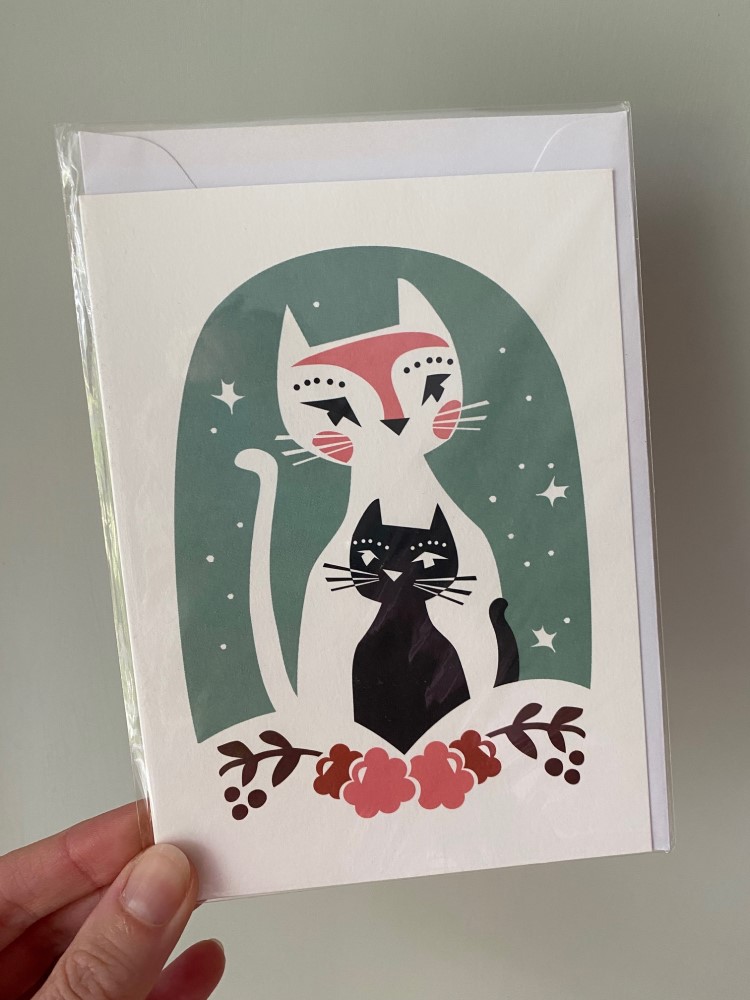 IMG_1940 Grußkarte, zwei stilisierte Katzen, eine weiß mit rosa Akzenten, eine schwarz, grüner Hintergrund, florale Elemente am unteren Rand, in transparenter Verpackung.