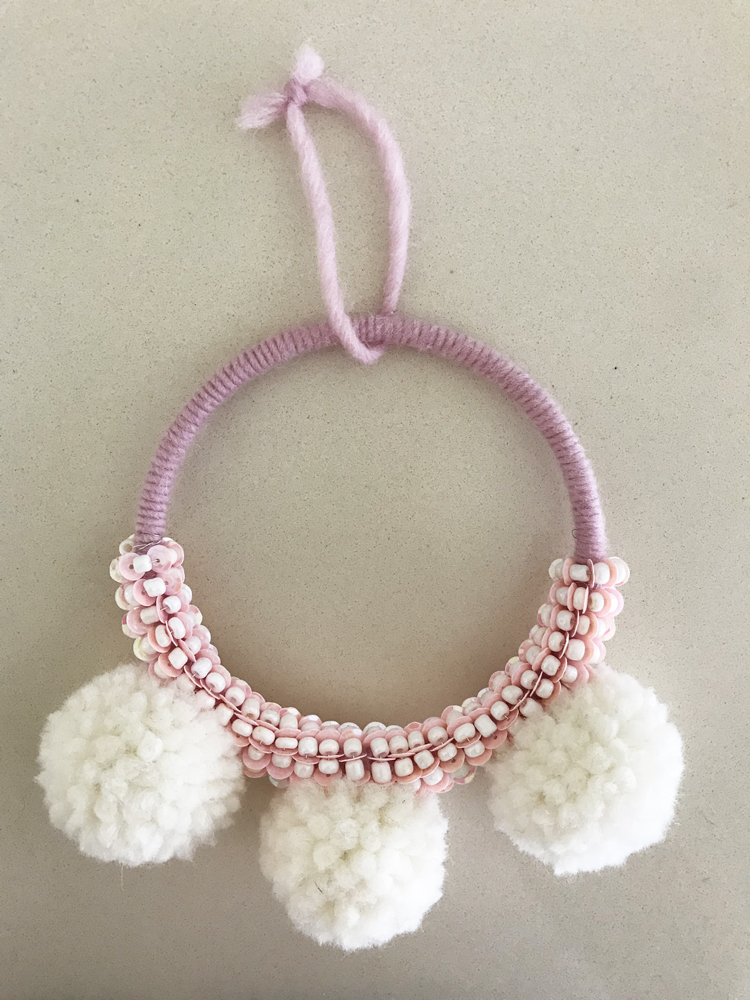 IMG_2697 Runder Wandbehang aus lilafarbenem Garn, verziert mit weißen und rosafarbenen Perlen, drei weiße Pompons an der Unterseite.