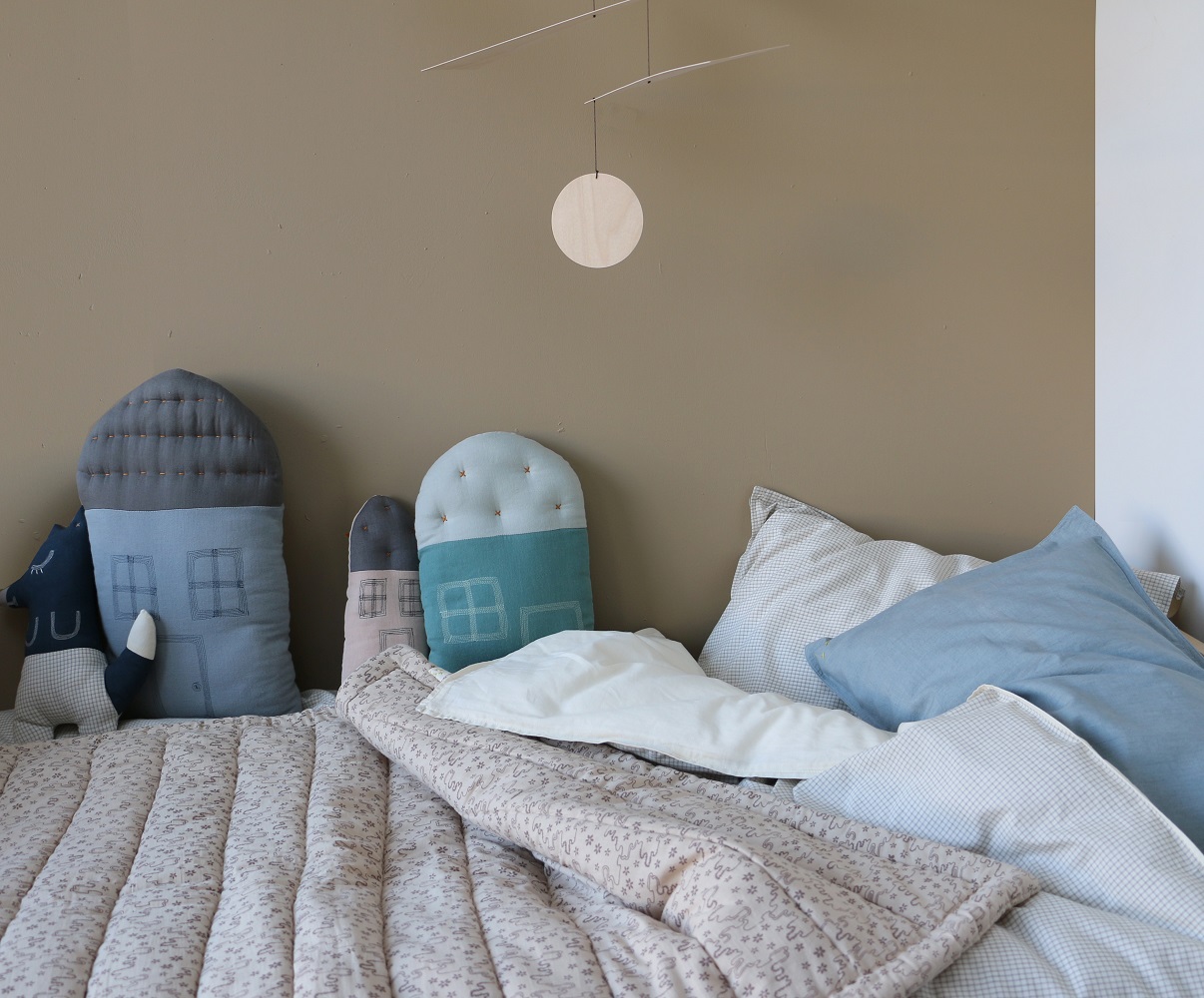 detail11 Kissen in Hausform, verschiedene Blautöne, gemusterte Bettdecke in Beige, Couch mit blau-weißen Kissen, Mobile aus Holz mit minimalistischem Design.