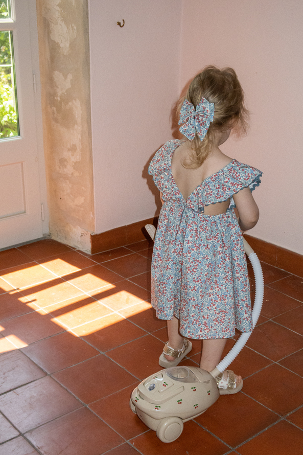 Kleinkind mit blonden Haaren, geblümtes Sommerkleid, passender Haarbogen, Miniaturstaubsauger-Spielzeug, beige mit kleinen bunten Motiven, Terrakottaboden.