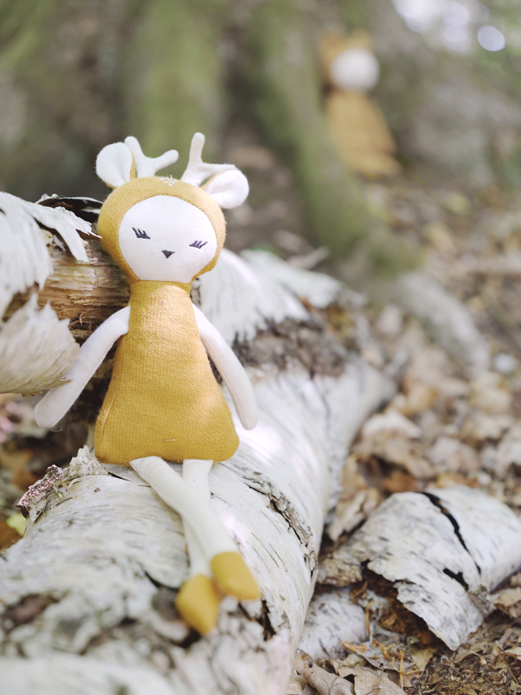 deer-on-log Stoffpuppe, Reh-Design, senfgelbes Kleid, cremeweiße Details, sitzend auf Baumstamm, kindliches Gesicht, geschlossene Augen, Ohren und Hörner.