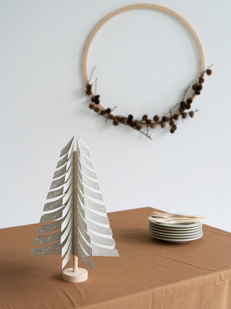 fir-tree-light-grey-4 Papier-Weihnachtsbaum, grau-weiß gemustert, auf Holzständer, auf braun gedecktem Tisch, daneben gestapelte weiße Teller mit Holzbesteck.