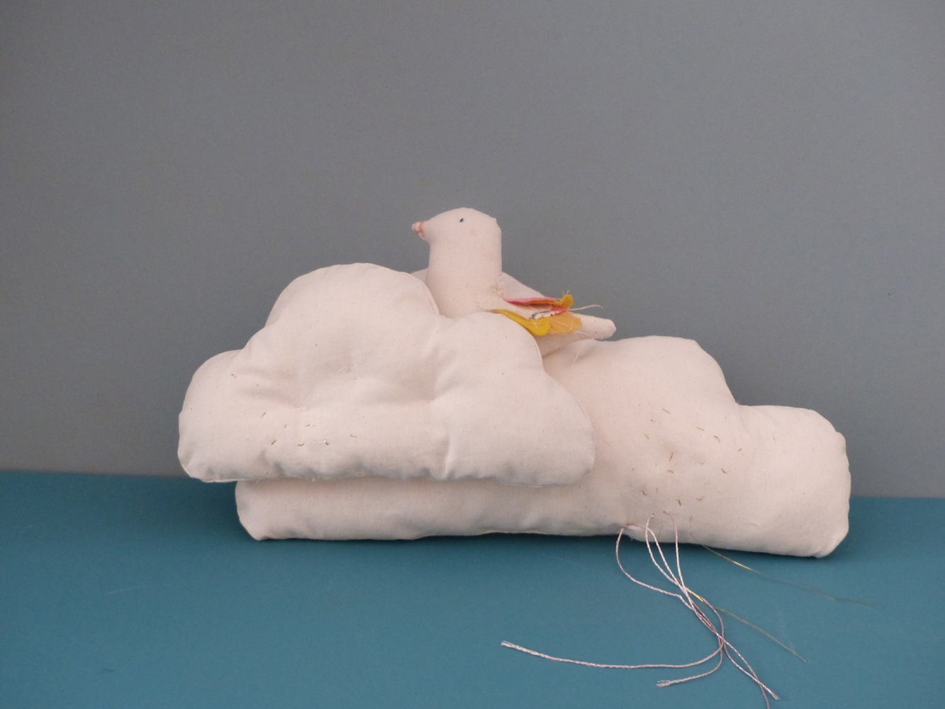 Scala-Suzie-nuage-2 Stofftier-Ensemble, weiße Wolke, darauf sitzender Vogel, pastellfarbene Flügel und Schwanzfedern, blaue Hintergrundfläche.