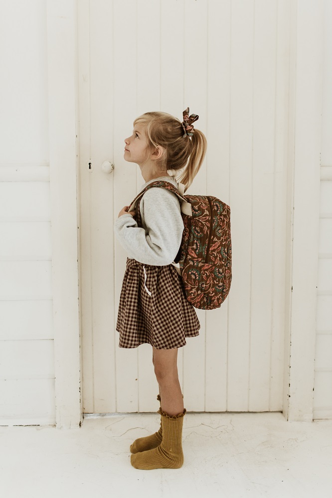 Girls-Backpack-Arti-BronzeFolkFlowers-1 Kind, blond, Pferdeschwanz, Haarband, grauer Pullover, karierter Rock, gelbe Socken, gemusterter Rucksack in Grün-Rot, Profilansicht.