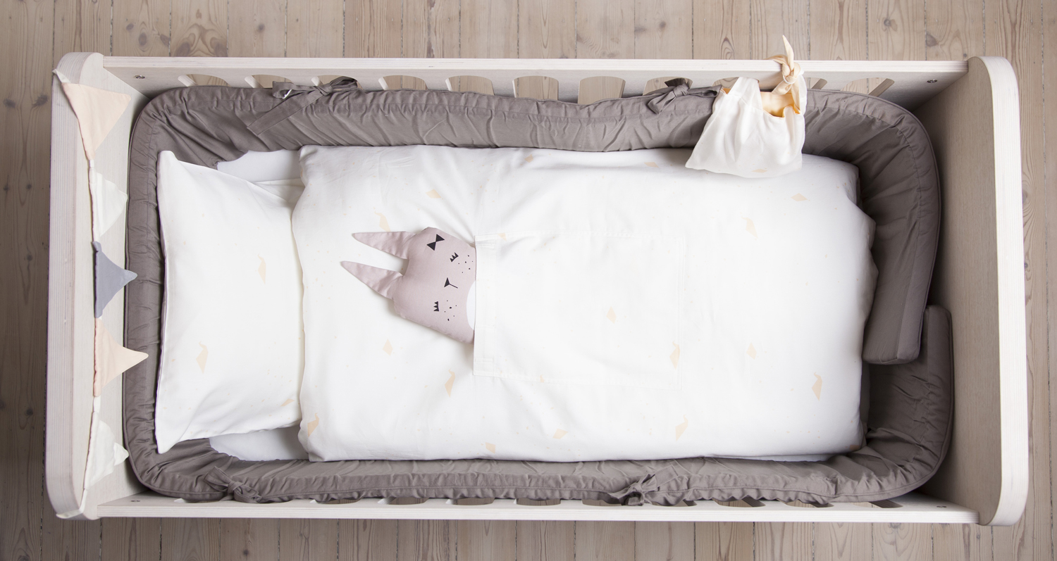 sb-BA16_baby-print-alisan-1 Babybett mit weißer Bettwäsche, grauer Umrandung, aufgedrucktem Tiergesicht im Bett, dreieckigen Stoff-Wimpeln in Creme, Grau und Beige am Rand.