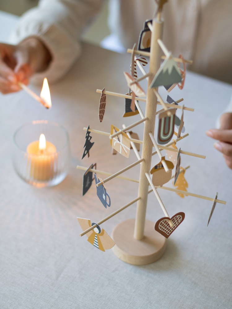 advent-tree-22 Holz-Weihnachtsbaum, minimalistisch, beige, Deko-Anhänger in verschiedenen Formen und Farben, darunter Herz, Tasse, Feder; Kerze im Glas, Personenhand mit Streichholz.