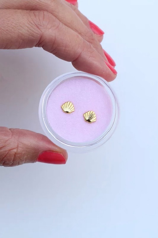 7a75ef_b8393a18c98b4bf0905b9d86d2804303_mv2 Mini-Goldmuschelohrringe, rundes durchsichtiges Etui, rosa Hintergrund, Hand mit rotem Nagellack.