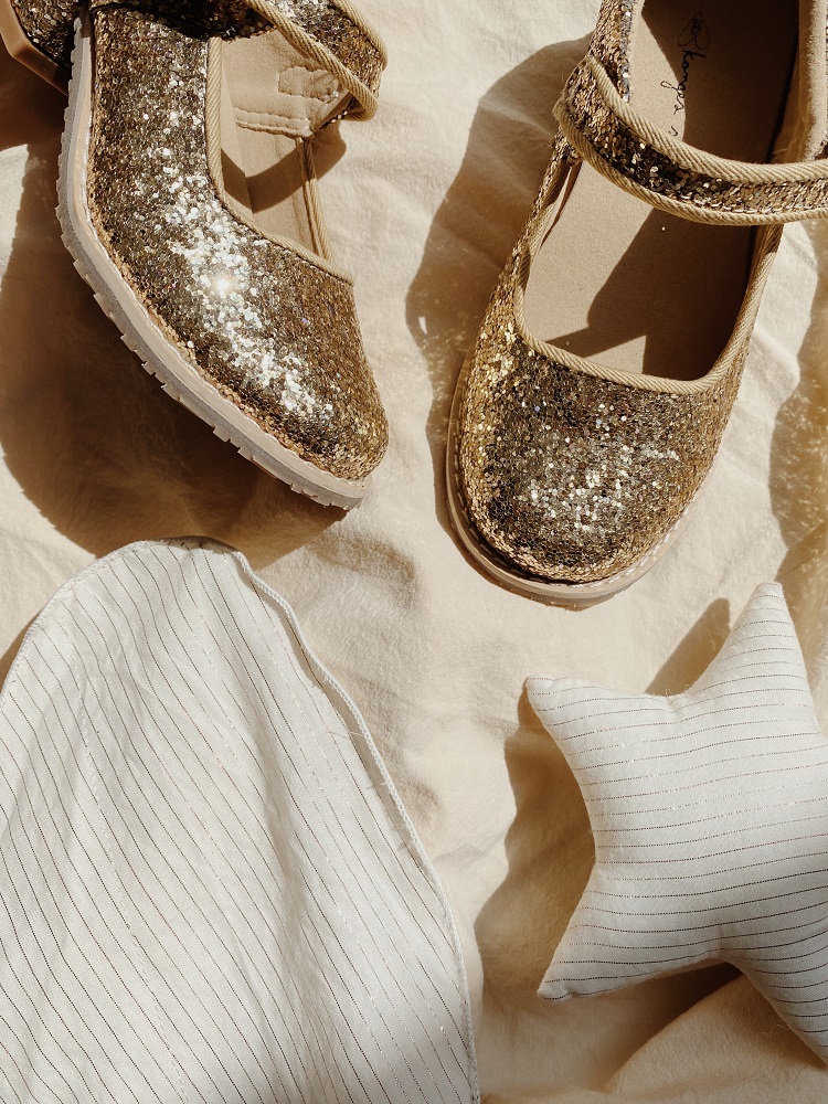KS1933-MULTI-Extra-2 Goldene glitzernde Ballerinas mit Riemen, weiße Sohle; daneben weißer Sternkissen und gestreiftes Stoffdetail.