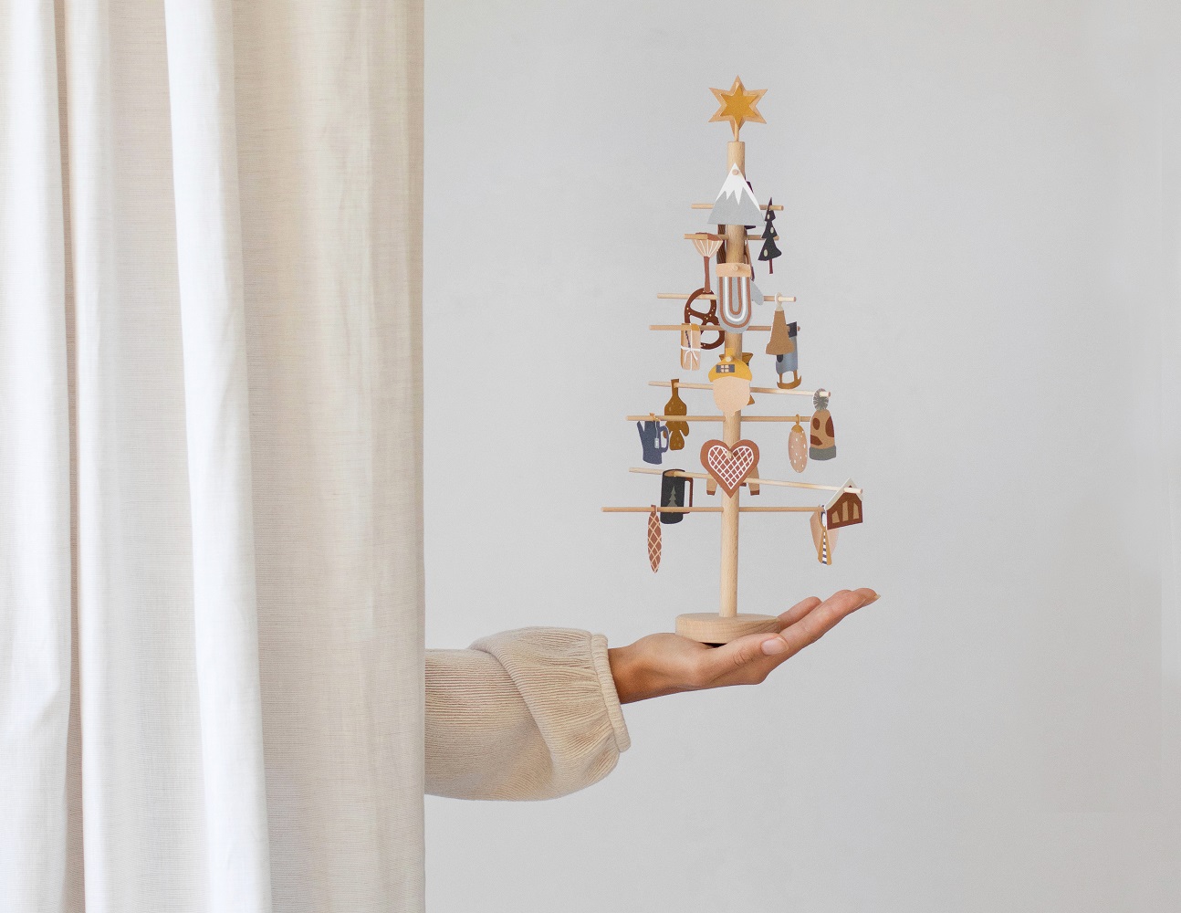 advent-tree-24ia9kcFhjcvPrw Holz-Weihnachtsbaum, Naturfarben, kleiner Stern oben, diverse miniaturartige Holzanhänger in Rot, Blau, Braun, Formen wie Herz, Haus, Mütze, aufrecht in Hand gehalten.