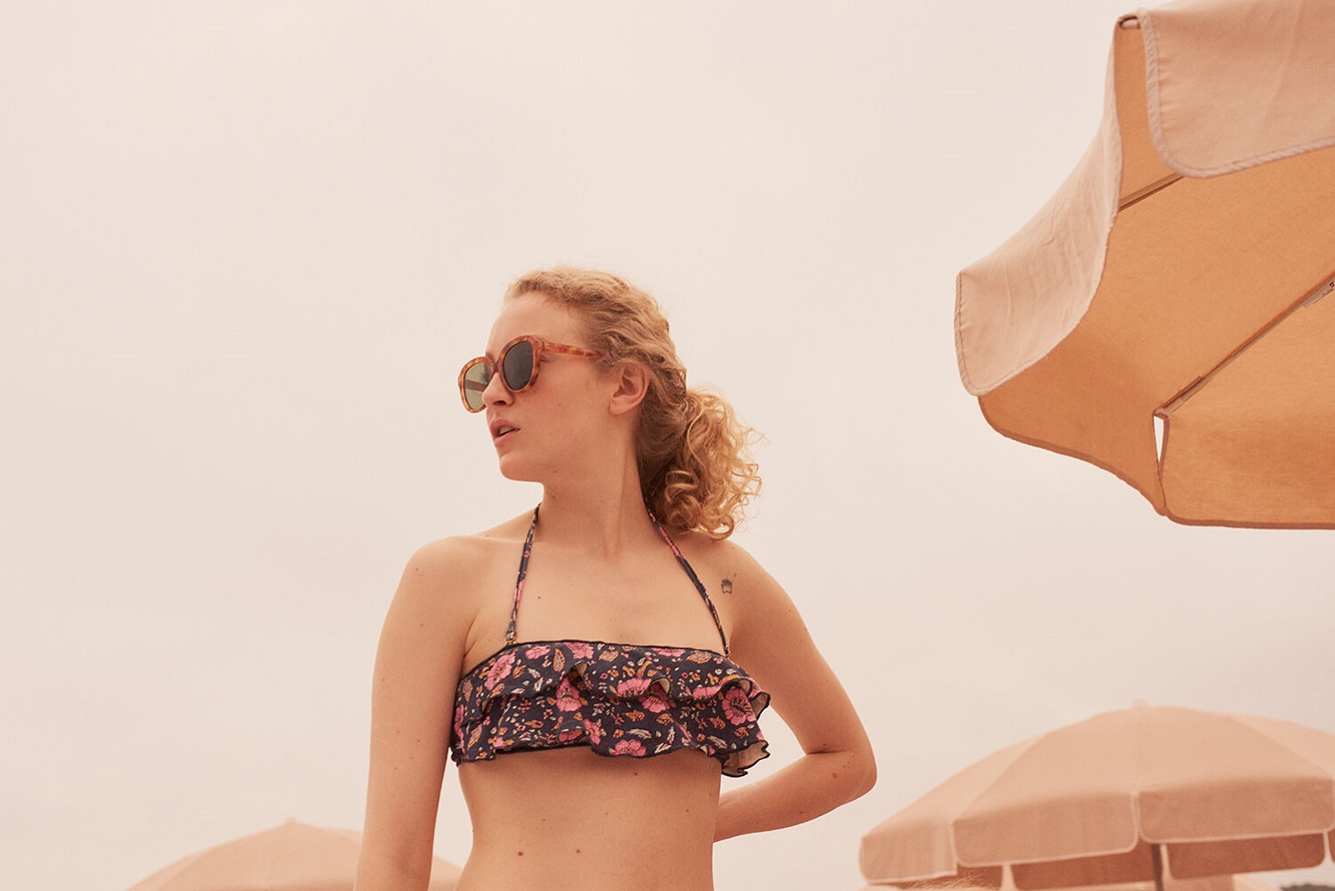 women-bikini-top-josia-black-flowers-1 Bandeau-Bikini, dunkel mit buntem Blumenmuster, Rüschen-Details, Sonnenbrille, lockiges Haar.
