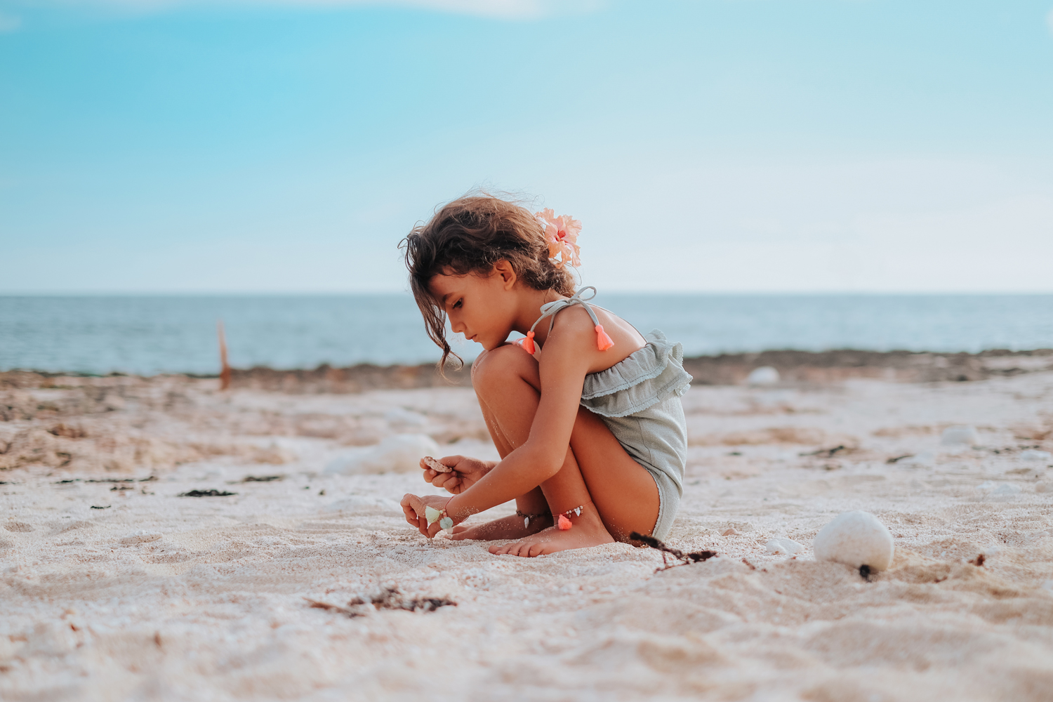 kidsbathingsuitcreolealmond11 Mädchen im grauen Badeanzug mit neonorangenen Details, sitzend am Strand, Muschel in der Hand, Meer im Hintergrund.