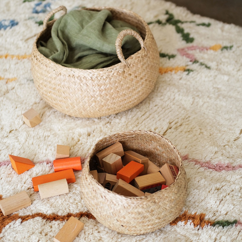 Olli-Ella-Robbie-Display-Bowl-Small-Large-Lifestyle-01-medium Zwei runde, geflochtene Körbe aus Naturmaterialien; großer Korb mit grünem Stoff; kleiner Korb mit Holzbausteinen in Natur- und Orangetönen; Korbgriffe; Teppich im Hintergrund.