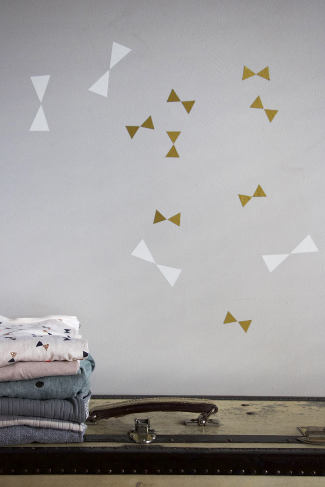 wallsticker-bows Gestapelte Kleidung, Farben: rosa, blau, grau, Muster: Dreiecke, Hintergrund: Wandsticker in Gold und Weiß, Kofferoberfläche als Unterlage.