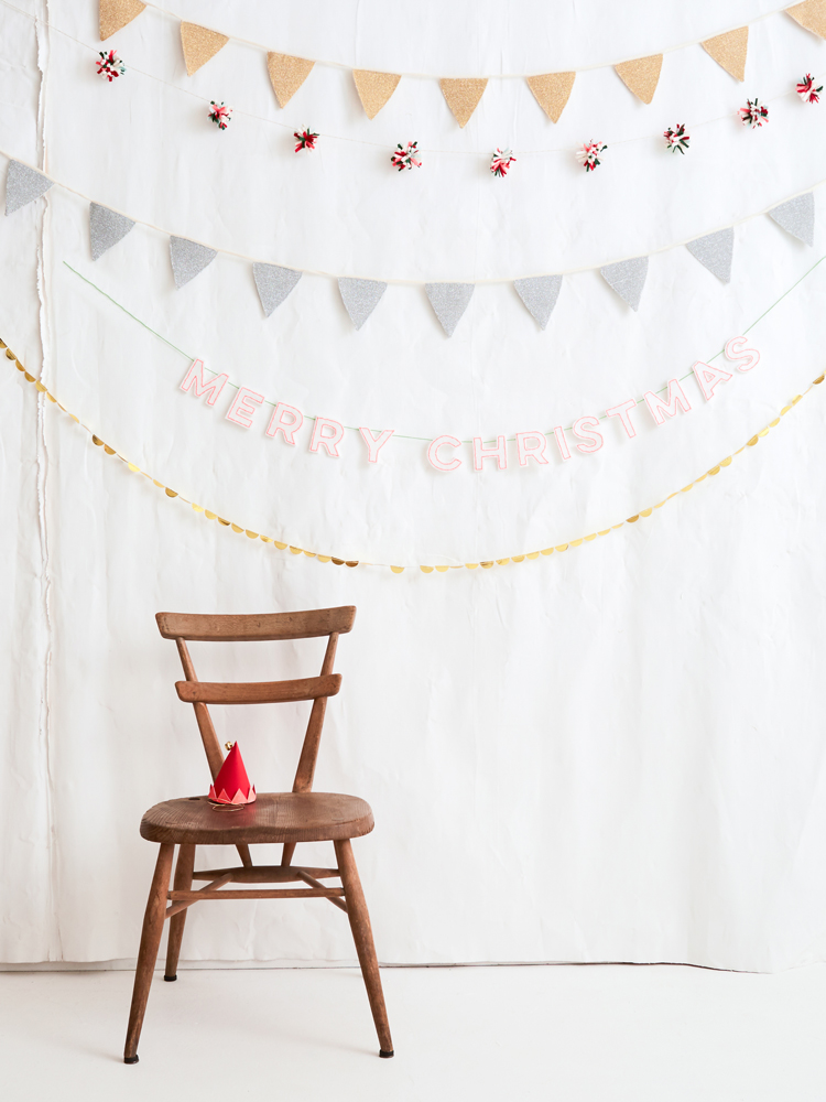 AW2017-HOME-4 Holzstuhl, rot-goldene Partyhütchen. Hintergrund mit Wimpelketten in Beige und Grau, Schriftzug "Merry Christmas" in Rosa, kleine rote und weiße Blumendekoration.