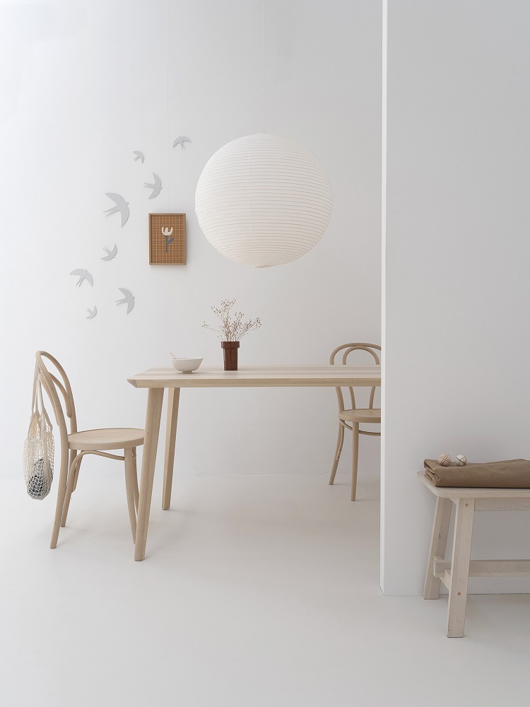 sverm-birds-large-2 Holztisch und zwei Holzstühle in hellem Naturton, runde weiße Papierlampe, weiße Wand mit Schwalben-Design, kleiner Holzrahmen mit Blume, Bank mit Decke.