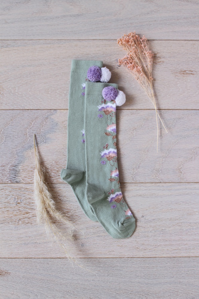 kidssocksmayumilichen1 Grüne Socken, Blumenmuster in Lila und Weiß, Pompons in Lila und Weiß.