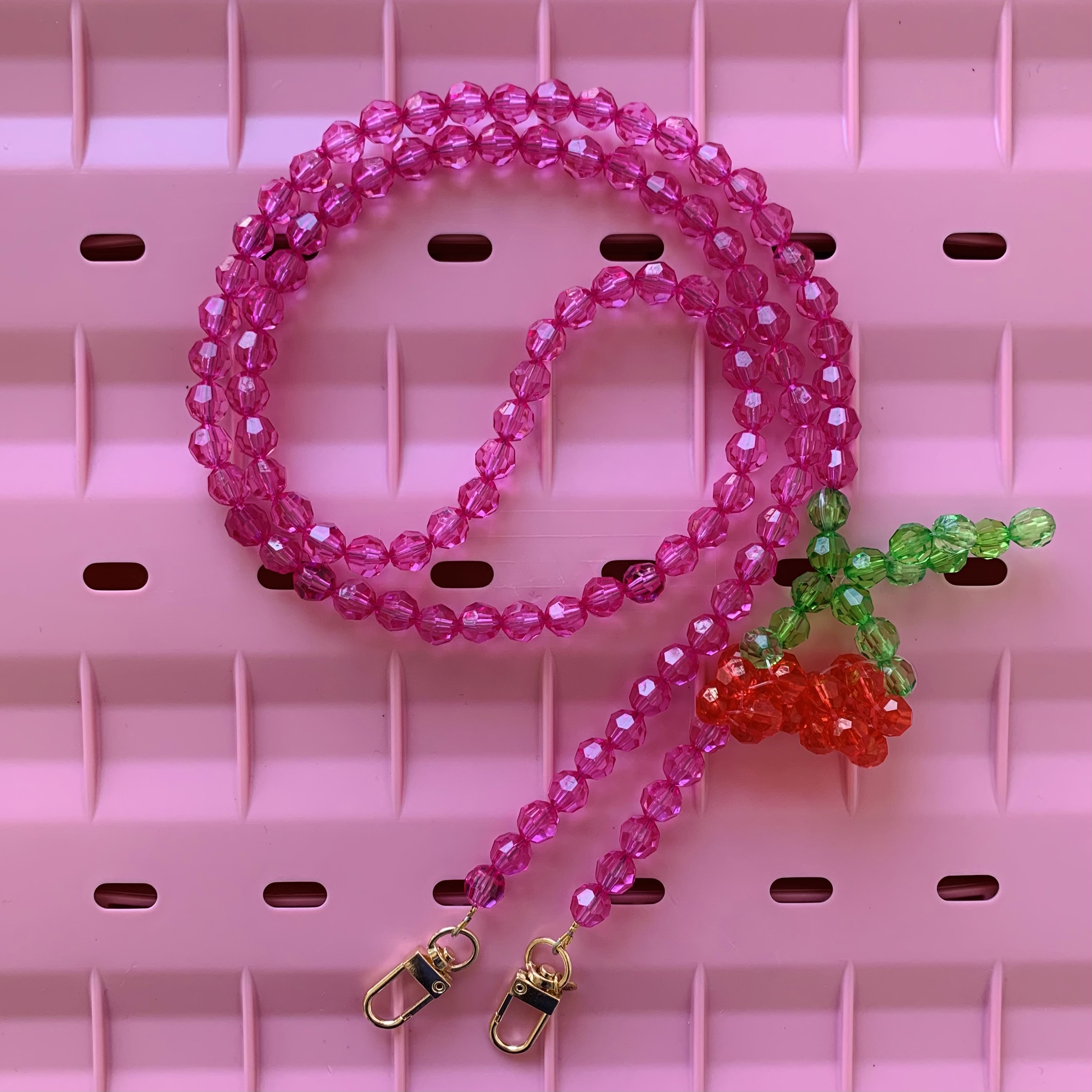 Perlenkette, pink, rote und grüne Perlen, Kirschenform, goldene Verschlüsse, rosa Hintergrund.