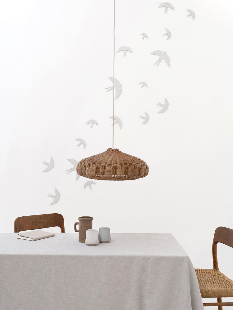 sverm-birds-large-3 Hängeleuchte aus Rattan, braun; Tisch mit beiger Tischdecke; zwei Holzstühle; Wanddekoration mit weißen Vogelsilhouetten; Keramiktassen und -krug auf dem Tisch.