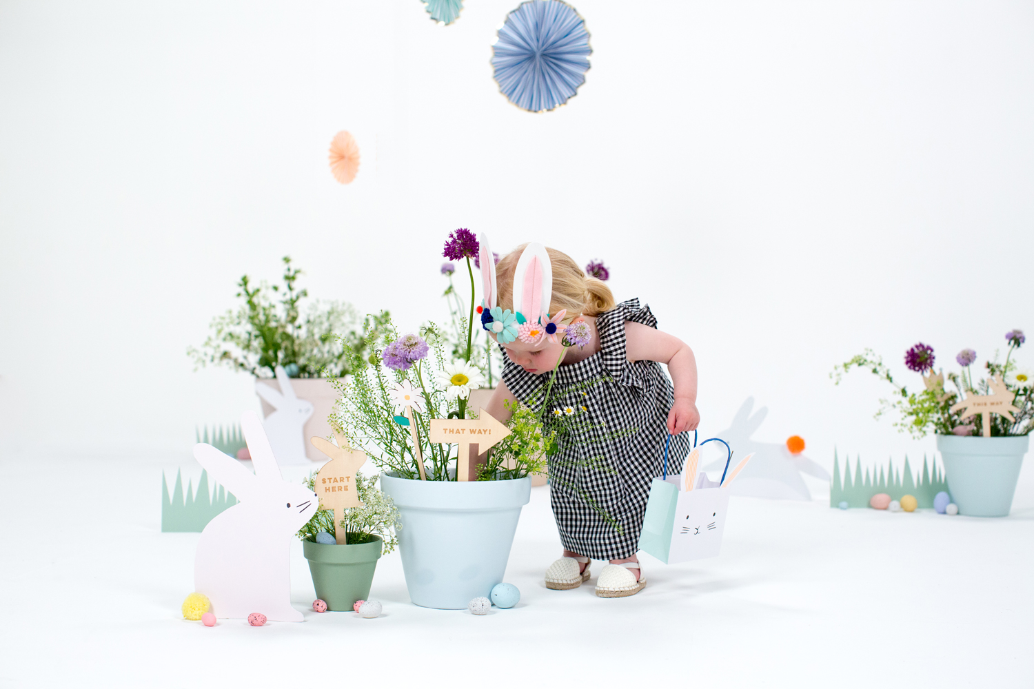 MM2018_Spring_Easter_12 Kind in schwarz-weiß kariertem Kleid, bunte Blumenkrone mit Hasenohren, weiße Sandalen; hellblaue Geschenktüte mit Hasenmotiv; Blumenkästen in Pastellfarben; Papierelemente wie Hasen und Schilder mit Aufdrucken.