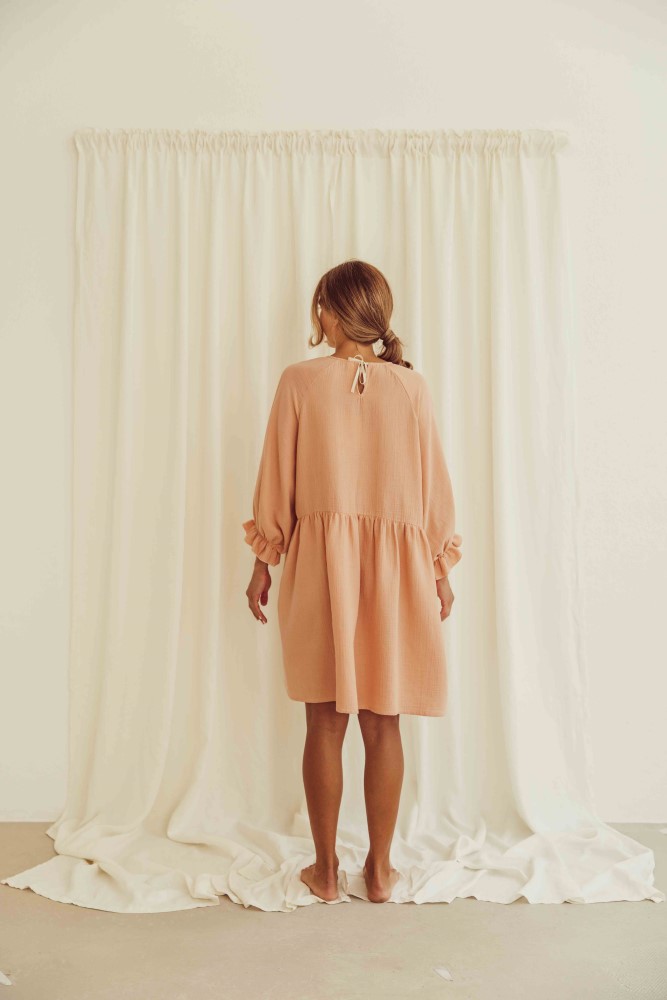 liss23_075_W_lilou-dress_peach_back Rückansicht Kleid, locker geschnitten, knielang, lange Ärmel, hellbraun, Bindeband am Rücken, Vorhang im Hintergrund.