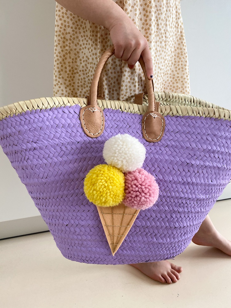 IMG_2832 Geflochtene Tasche, lila Farbe, Eiscreme-Pompon-Aufdruck, gelb-weiß-rosa, braune Tragegriffe, beige-weißes Kleid im Hintergrund.