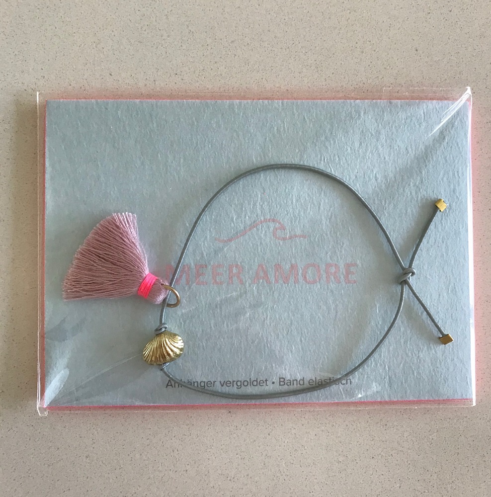 IMG_5903 Elastisches Armband, graues Band, vergoldeter Muschel-Anhänger, rosa Quaste, Papierhintergrund mit Aufdruck "MEER AMORE".