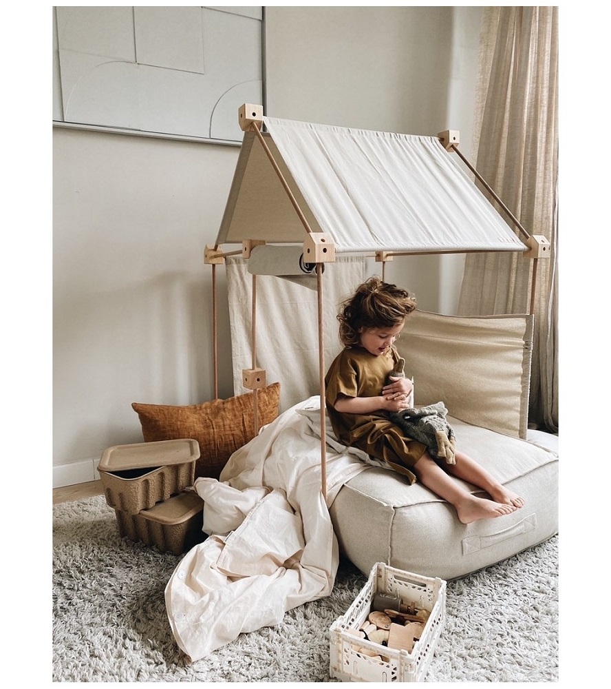 lniany-puf-podlogowy-naturalny-bez-3 Kind auf Sitzsack, Spielhaus aus Holz und Stoff, beige Decken, Aufbewahrungskisten, Plüschspielzeug in Tierform, natürliche Farbtöne.