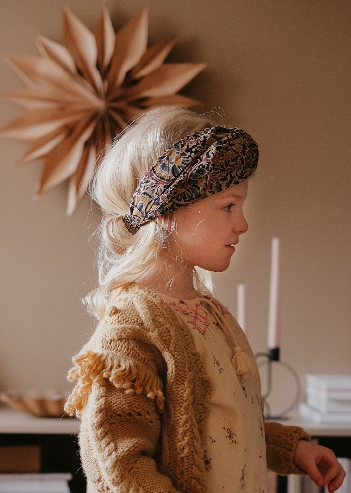 Girls-Headband-Daia-NordishFlowers-5 Kind trägt Stirnband mit floralem Muster; Kleid mit feinem Blumenaufdruck; gestrickte Jacke, beige mit Fransen.