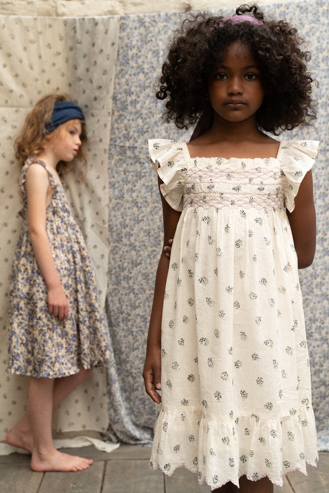 _DSC6249 Mädchenkleid, weiß, Stoffmuster mit Blumen, Rüschen an den Ärmeln, verspielter Saum. Im Hintergrund weiteres Mädchen mit blauem Blumenmuster-Kleid, Haarband.