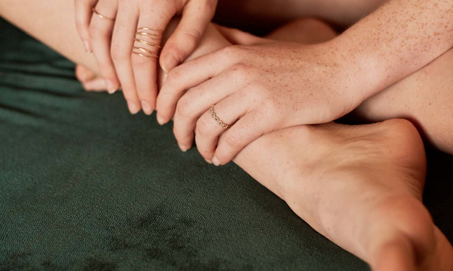 women-ring-carlett-gold-2 Personenhände mit goldenen Ringen, liegend auf dunkelgrünem Stoff, Fokus auf Schmuck.