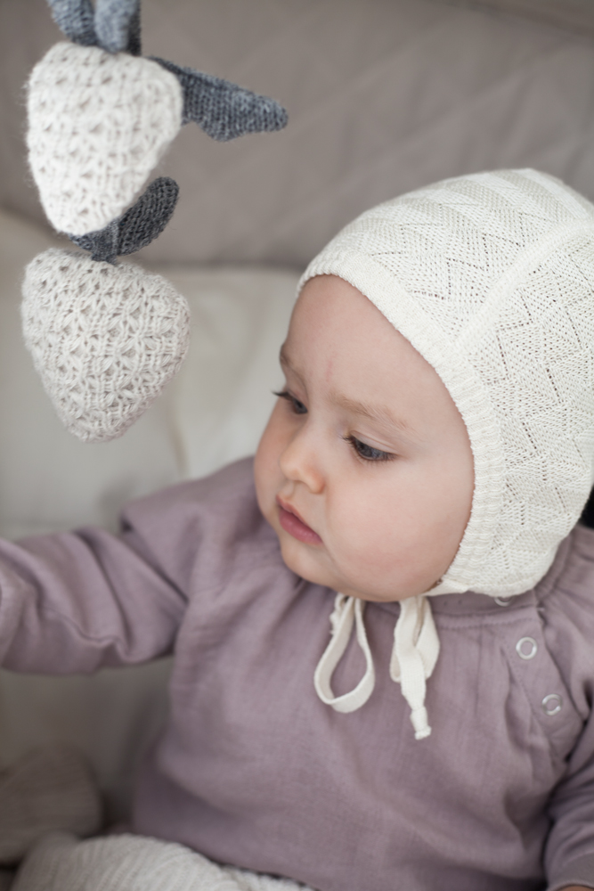 57 Baby, cremeweiße gestrickte Mütze, lila Oberteil, gestricktes Mobile in Herzform, weiß und grau.