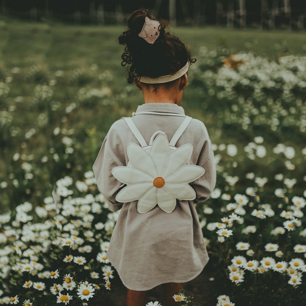 5006003_LE060_2 Kinder-Rucksack, Blütenform, Weiß mit gelbem Zentrum, auf dem Rücken eines Kindes, helles Kleid, Blumenwiese im Hintergrund.