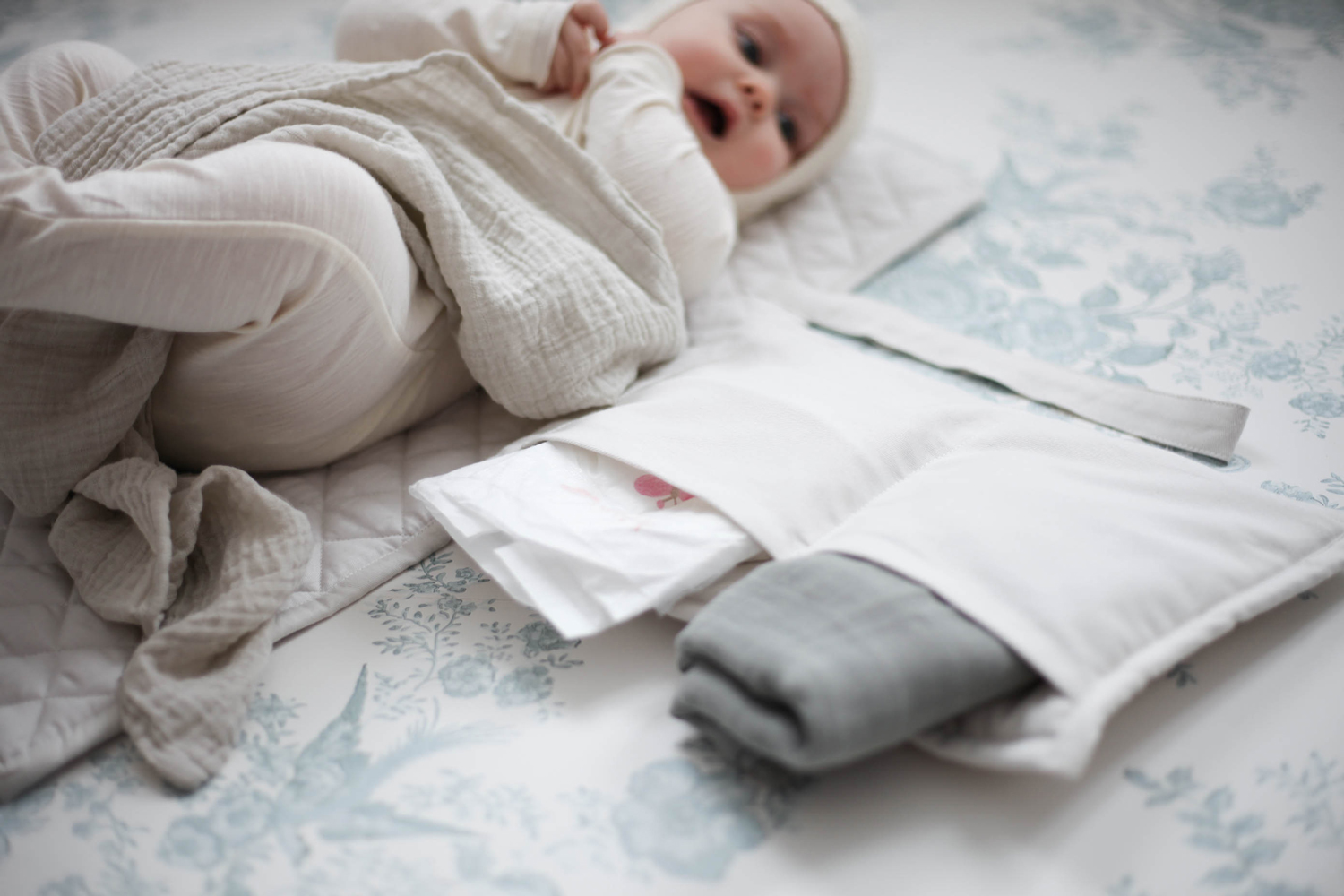 IMG_7109-2 Babyausstattung, hellbeige Wickelunterlage, weißes Tuch, graues Stoffbündel, Baby in weißem Strampler auf gemusterter Decke.