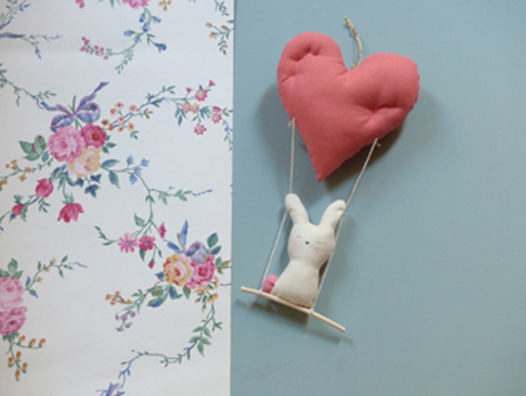 coeur-et-lapin2 Tapetenmuster mit Blumen, pastellfarben; Stoffherz-Aufhänger in Rosa; Hänge-Wippe mit weißem Stoffkaninchen, sitzend.