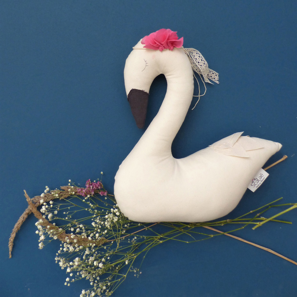 Scala-Swan-le-cygne-4 Stofftier Schwan, cremeweiß, schwarzer Schnabel, rosa Stoffblume am Kopf, Spitze und Schnur als Dekoration, blauer Hintergrund.