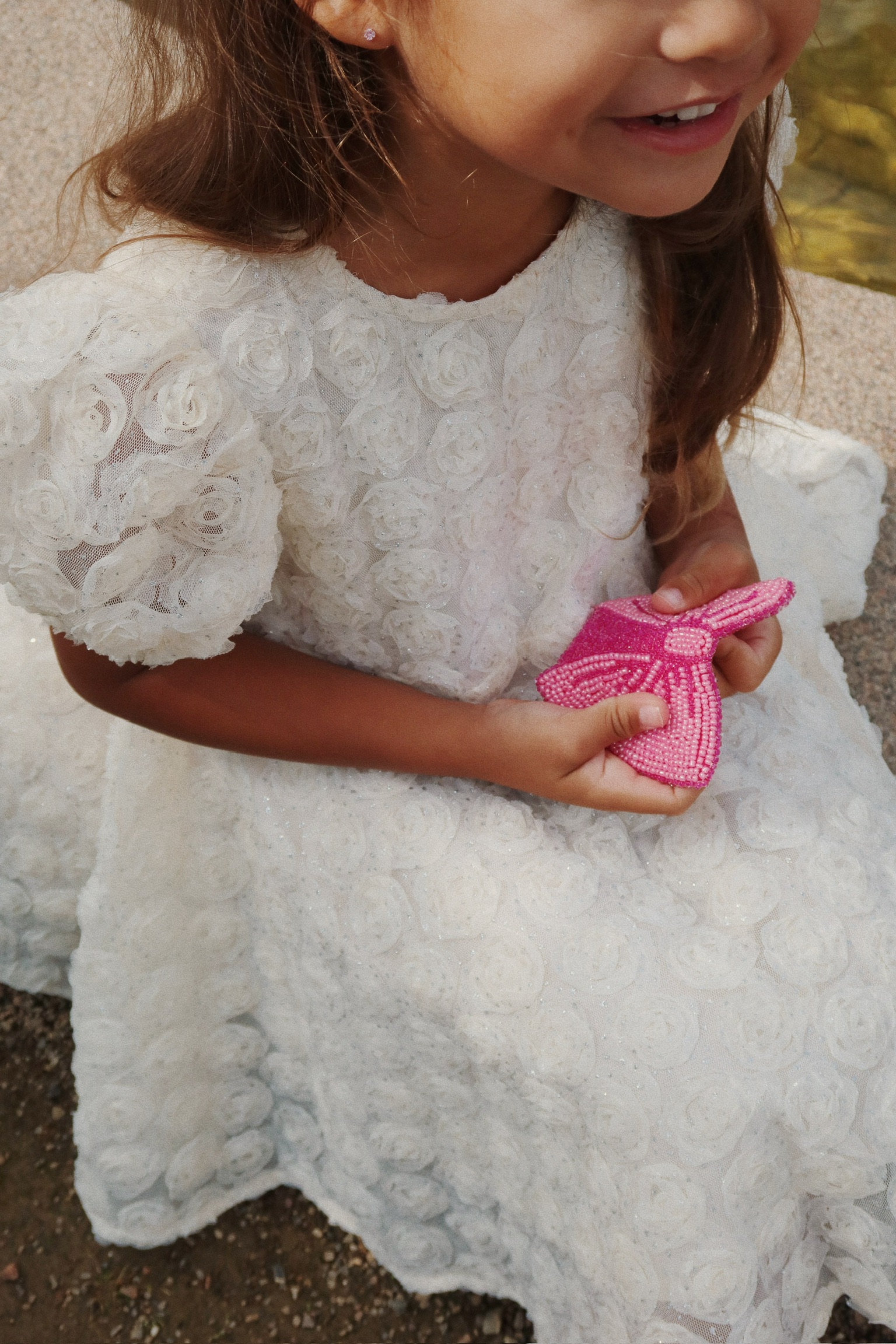 Kind in weißem Spitzenkleid mit Blumenmuster, hält pinke, glitzernde Perlen-Clutch in Schmetterlingsform.