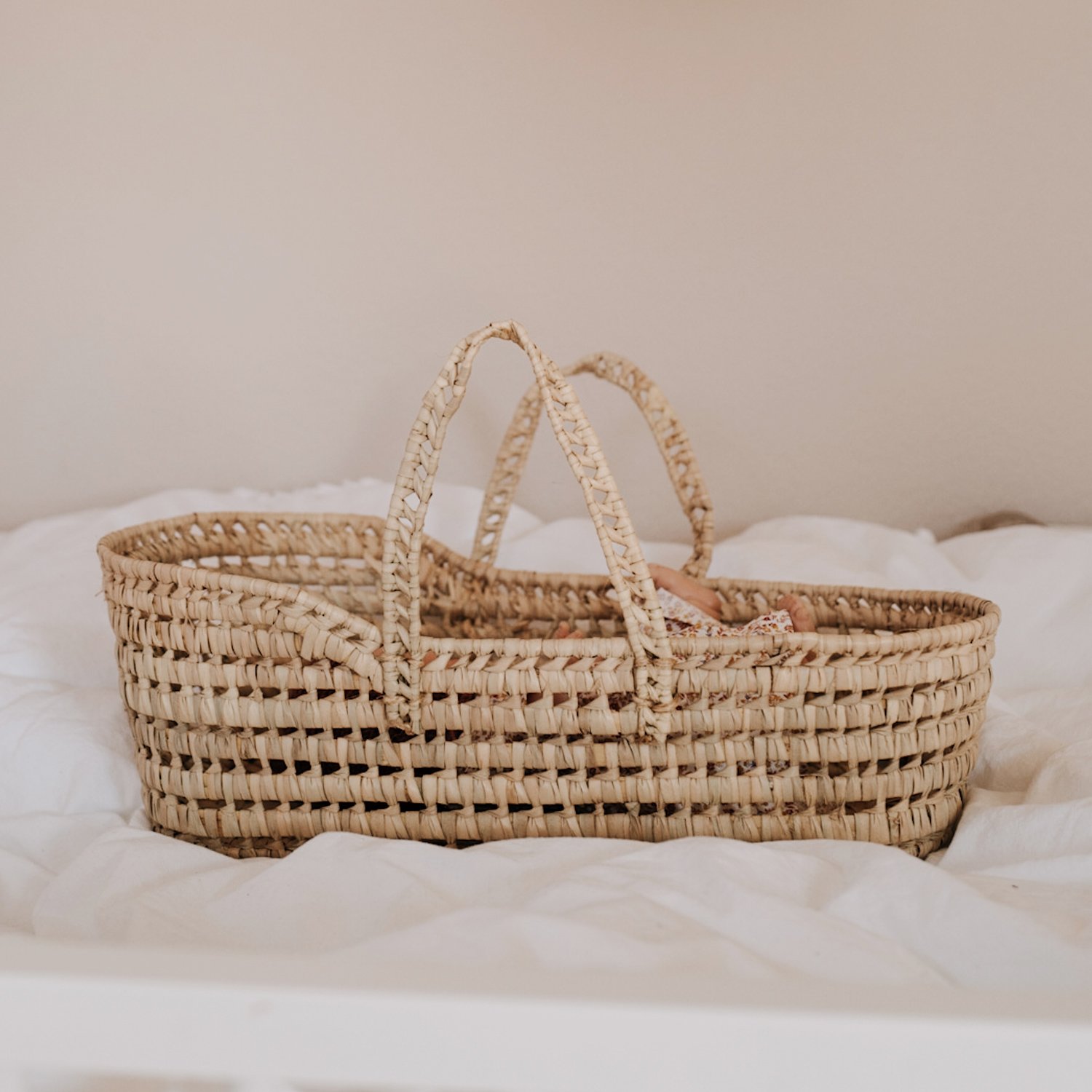 lucylou_puppentrage_02_1024x1024-2x Geflochtener Baby-Moseskorb, beige, zwei Henkel, auf weißem Bett.