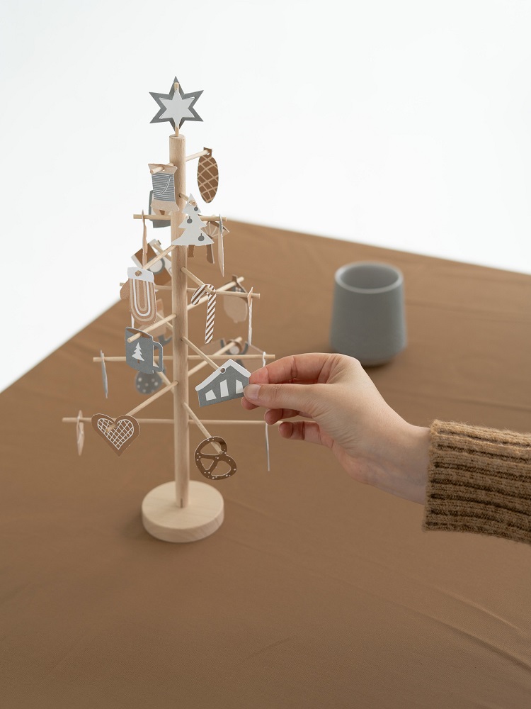 advent-tree-natural-3 Holz-Weihnachtsbaum, naturbelassen, abnehmbarer Schmuck; weiße und graue Anhänger: Stern, Baum, Herz, Haus, Rentier; Arm im Strickpulli hält Haus-Anhänger; Tisch mit beigem Tuch.