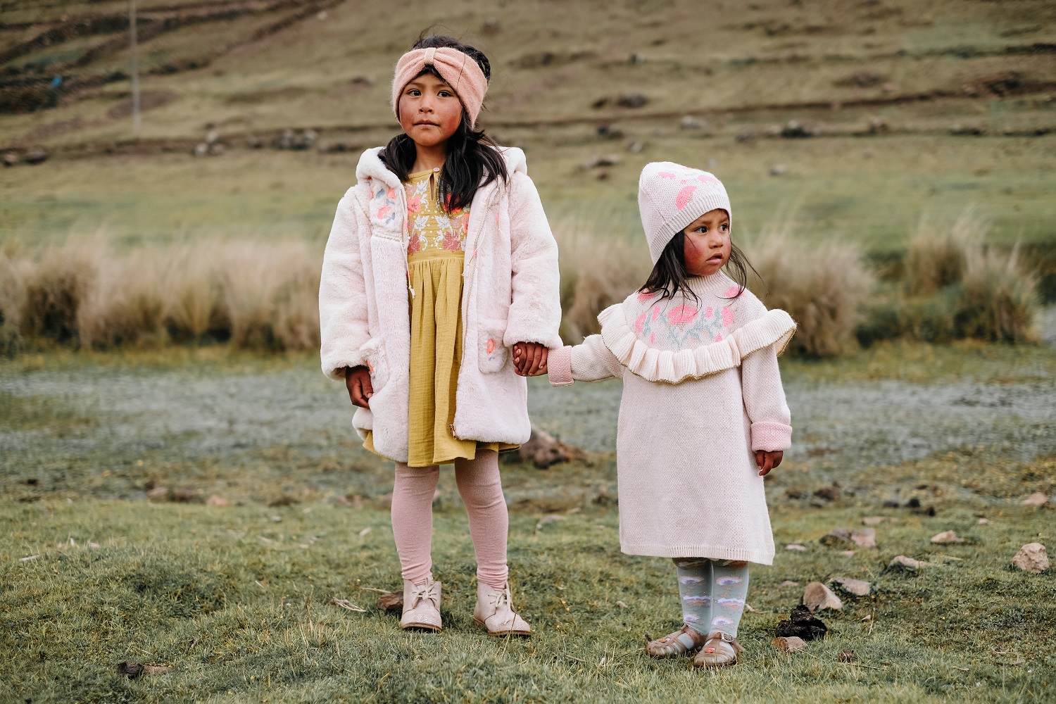 kidssocksmayumilichen3 Zwei Kinder, eine mit rosa Teddyjacke, gelbem Kleid, rosa Stirnband; die andere mit rosa Strickkleid, passenden Mütze, Strumpfhose mit Muster. Beide halten sich an den Händen, stehen auf Wiese. Kleidung farbig gemustert.