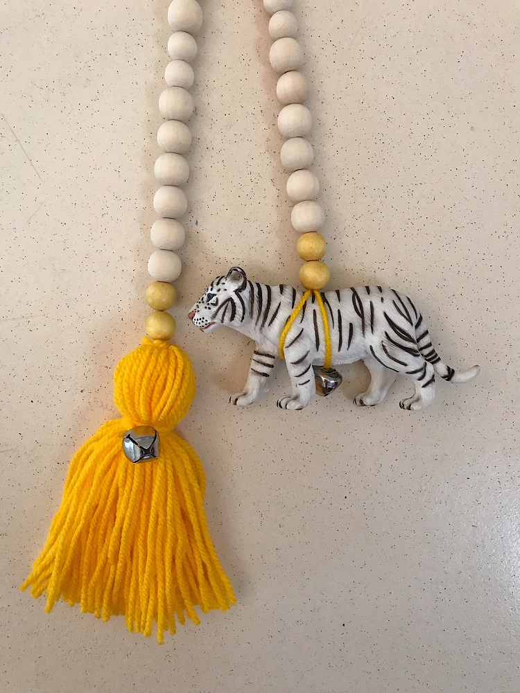 0FC69B2F-E444-4DFE-8283-41310F7B6AC9-002 Holzperlenkette, cremefarben, gelb; Tigerfigur, weiß-schwarz gestreift; gelber Wollquast; kleine Glocke.