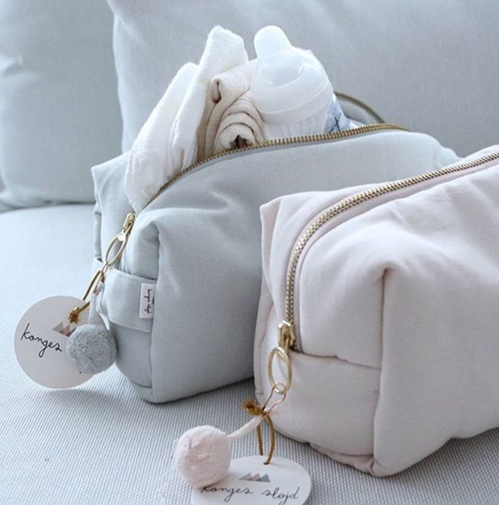 FullSizeRender Zwei Kosmetiktaschen, eine hellgrau, eine rosa, mit Reißverschluss, Pompon-Anhänger, Etikett "Konges Slojd"; hellgraue Tasche gefüllt mit Babyflasche und Stoffen.